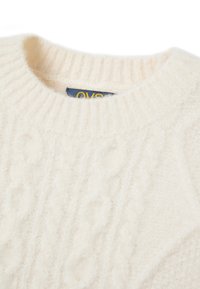 Cremfarbener, grob gestrickter Pullover mit rundem Halsausschnitt und gerippten Details. Der Stoff wirkt weich und strukturiert. Etikett am Hals sichtbar.