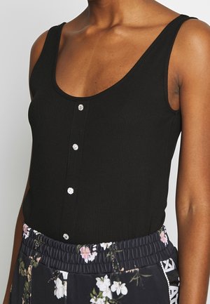 Femme portant un débardeur noir côtelé avec quatre petits boutons décoratifs, associé à un pantalon noir à motif floral avec taille élastique.