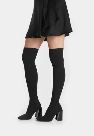 Bottes noires au-dessus du genou en tissu extensible, avec un bout pointu et un talon épais, dotées d'un zip sur le côté intérieur.