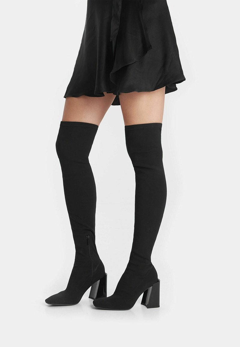 Botas negras por encima de la rodilla hechas de un tejido elástico, con punta afilada y tacón grueso, presentando un detalle de cremallera en el interior.