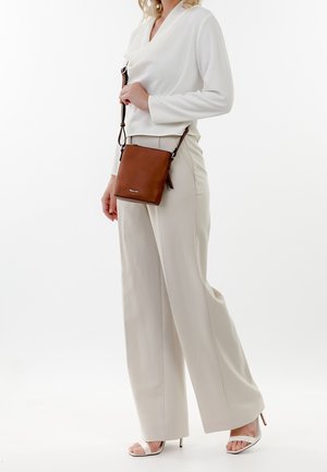 Kvinde i hvid bluse, beige vide bukser, hvide højhælede sandaler, der holder en brun crossbody lædertaske.