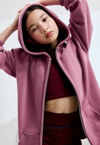 Ružová mikina na zips s prednými vreckami, mäkkou textúrou látky a podšitým kapucňou, ktorá je oblečená na bordovú crop top a zodpovedajúce legíny s vysokým pásom.