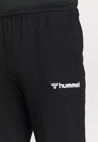 Pantalon de survêtement noir à taille élastique avec logo blanc "hummel" et emblème d'abeille sur la cuisse gauche, présenté sur un fond clair uni.
