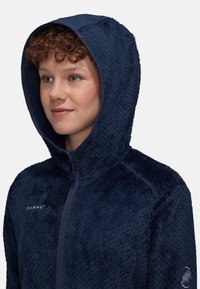 Mörkblå fleecejacka med huva, featuring en strukturerad yta. Den har hel dragkedja och logotypdetaljer på bröstet och ärmen.