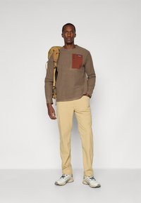 Brauner Strickpullover mit einer Brusttasche in kontrastierendem Rostton, beigen Hosen und grau-weißen Sportschuhen, getragen von einem männlichen Modell mit einem Rucksack.