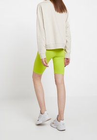 Shorts da bici aderenti di colore verde neon abbinati a una felpa sfusa color crema, indossati con scarpe da ginnastica bianche. Tessuto liscio con una texture lucida.