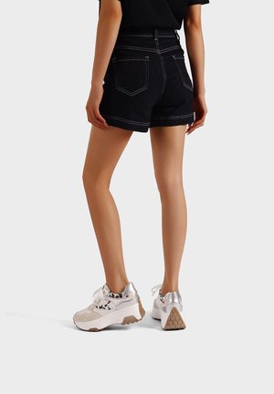 Persona che indossa pantaloncini di jeans neri e scarpe da ginnastica bianche con dettagli leopardati, in piedi davanti a uno sfondo grigio uniforme.