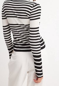 Personne portant un haut à manches longues côtelé à rayures horizontales noires et blanches, associé à un pantalon blanc taille haute, vue de dos.