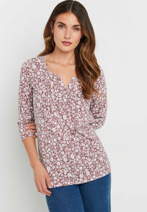 Bloes met bloemenprint in stofroze met witte bloemen, V-hals, driekwart mouwen en geplooide details. Gecombineerd met blauwe denim jeans.