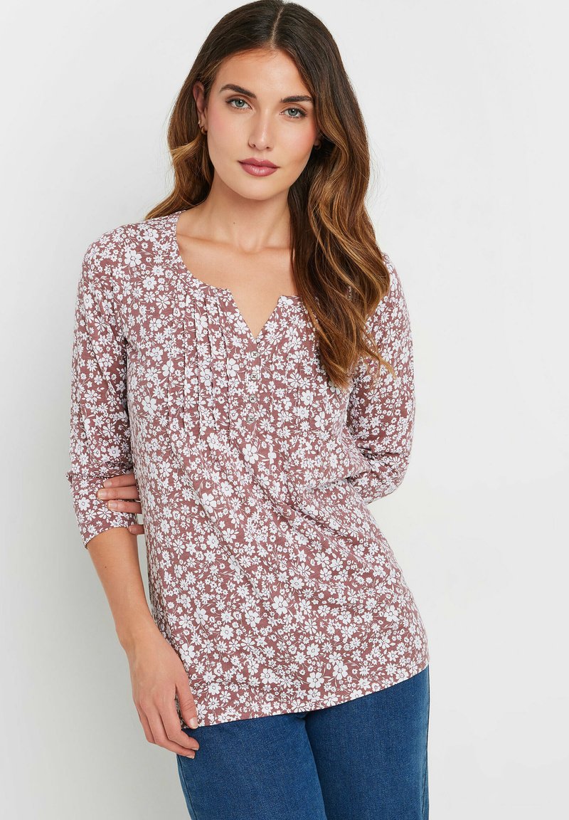 Bluse mit floralen Druck in staubigem Rosenfarben und weißen Blumen, V-Ausschnitt, drei Viertel Ärmel und gefaltete Details. Kombiniert mit blauen Jeans.