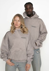 Grå huvtröjor gjorda av mjukt tyg med en känguruficka, avslappnad passform och "Multiply Apparel"-logotyp på bröstet. Bärs tillsammans med ljusa jeans.