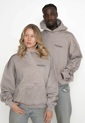 Sivé mikiny vyrobené z mäkkej tkaniny s kangaroo vreckom, voľný strih a logo "Multiply Apparel" na hrudi. Nosené s svetlými džínsy.