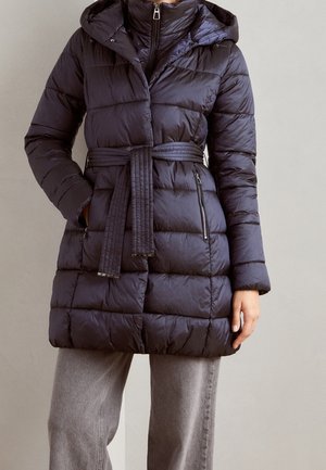 Personne portant un manteau matelassé noir mi-long avec capuche, taille ceinturée, poches zippées et pantalon gris.