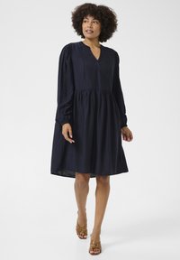 Vestido azul marino de manga larga con escote en V, cintura fruncida y falda acampanada. Hecho de una tela suave con una textura lisa. Sandalias de tacón marrones.