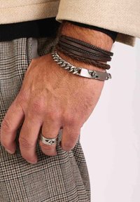 Lederen en metalen armbanden gestapeld op een pols, met gevlochten bruin leer, zilveren kettingschakels en een zilveren ring met een abstract ontwerp.