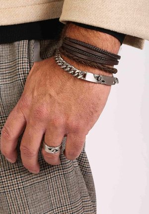 Lederen en metalen armbanden gestapeld op een pols, met gevlochten bruin leer, zilveren kettingschakels en een zilveren ring met een abstract ontwerp.