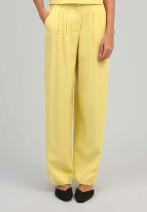 Femme portant un pantalon ample jaune pastel à plis sur le devant et des chaussures plates noires à bouts pointus, debout avec une main dans la poche.