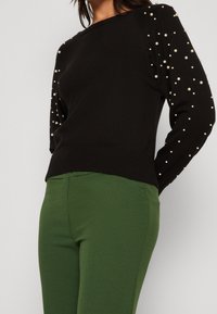 Schwarzer Pullover mit Perlenakzenten entlang der Ärmel, geripptem Saum, kombiniert mit grünen, eng anliegenden Hosen. Weiche Textur, Rundhalsausschnitt.
