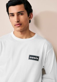 Balta ilgomis rankovėmis marškiniai, pagaminti iš medvilnės, su juodu stačiakampiu "oasis" logotipu kairėje krūtinės pusėje. Lygus paviršius, klasikinis siluetas.