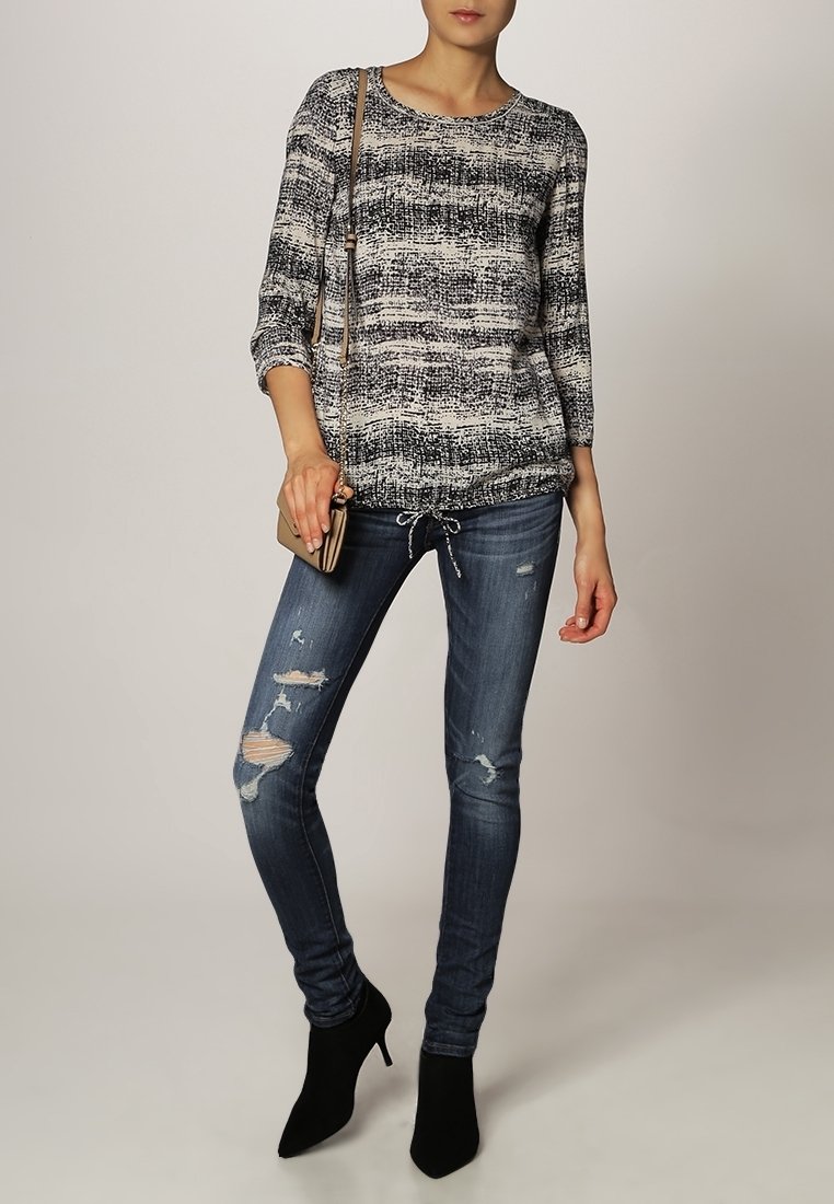 Femme portant un haut à manches longues noir et blanc à motifs, un jean skinny déchiré, des bottines noires et tenant un petit sac beige.