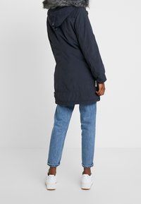 Navy parka with a fur-lined hood, featuring a slightly loose fit and elastic cuffs. Paired with light blue jeans and white sneakers.