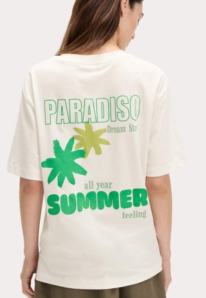 Person iført hvid, oversize T-shirt med grønne palmebladsgrafikker og tekst, der læser "PARADISO," "Dream Sta" og "all year SUMMER feeling."