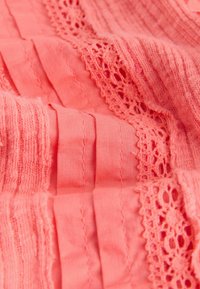 Tissu rose corail avec plis, parties en maille texturée et bordure en dentelle florale créant un motif textile superposé.