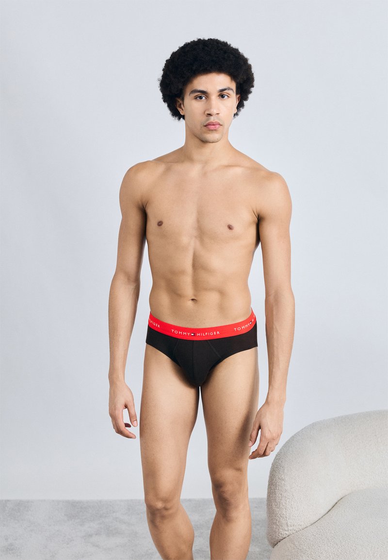 Μαύρο εσώρουχο σλιπ με κόκκινη ελαστική μέση που φέρει το λογότυπο "TOMMY HILFIGER". Το μοντέλο στέκεται μπροστά σε ουδέτερο φόντο.
