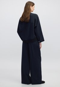 Chaqueta oversized de satén negro con mangas largas, dobladillo acanalado y una costura trasera sencilla, combinada con pantalones de pierna ancha de la misma tela.