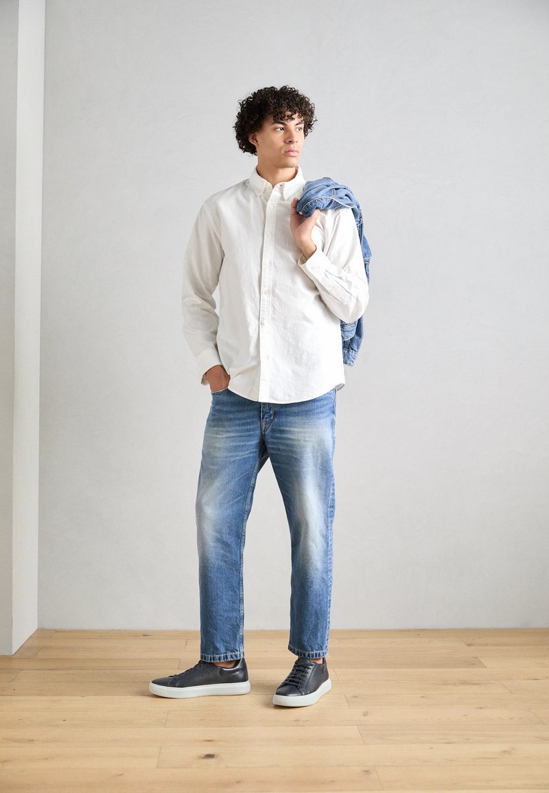 Chemise blanche à boutons, jeans bleu clair avec décoloration, baskets noires avec semelles blanches, et une veste en denim portée sur l'épaule.