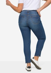 Jeans de mezclilla azules ajustados con cintura alta, dos bolsillos traseros y un dobladillo de tobillo ligeramente deshilachado, combinados con zapatillas blancas.