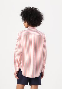 White shirt with red vertical stripes, buttoned cuffs, and a rounded hem. Back view shows a collar and placket with a single button accent.