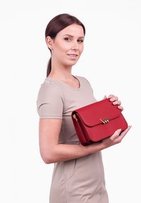 Červená kožená kabelka s přední klopou a zlatými kovovými doplňky. Kabelka má strukturovaný design a drží ji osoba oblečená v béžových šatech.