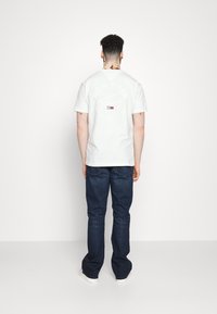 Camisa blanca de manga corta con un cuello redondo y un estampado gráfico en la parte posterior. Combinada con jeans azul oscuro y zapatillas blancas.