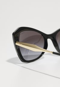 Óculos de sol grandes em forma de olho de gato, pretos, com lentes cinzentas em degradé e um detalhe em metal dourado na haste. Armação de plástico suave com ângulos marcantes.
