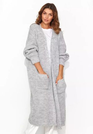 Kobieta w długim, grubym, szarym swetrze rozpinanym z dużymi przednimi kieszeniami, podwiniętymi rękawami i białym ubraniem pod spodem, stojąca na białym tle.