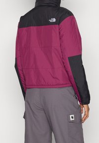 Lilla og sort dunjakke med et kort, tætsiddende design. Har en ståkrave, elastiske manchetter og The North Face-logoet på ryggen.