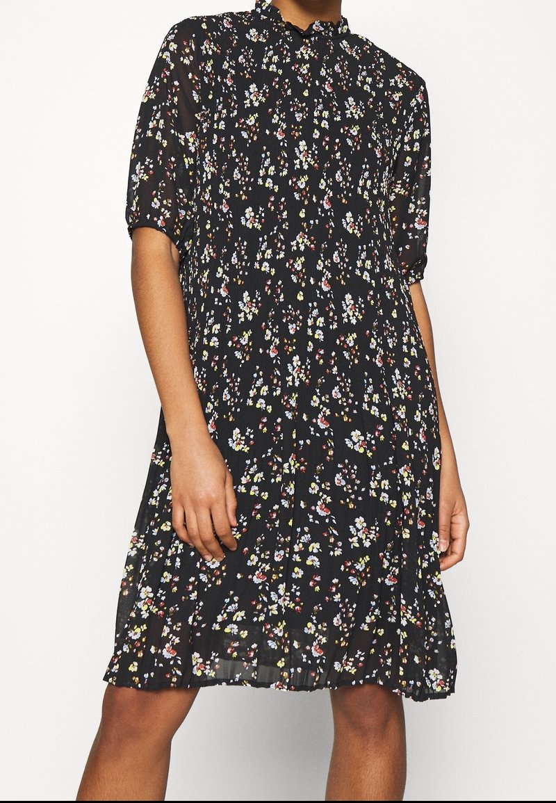 Robe noire longueur genoux avec un petit motif floral blanc, jaune et rouge, manches courtes bouffantes et col montant à volants.