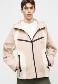 Νεαρός άνδρας που φορά μπεζ και λευκό φούτερ με κουκούλα και φερμουάρ, με μαύρες λεπτομέρειες, στέκεται με τα χέρια στις τσέπες πάνω σε απλό φόντο.