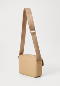 Liten rektangulær beige crossbody-veske med justerbar bred stoffrem og hardware i sølvmetall.