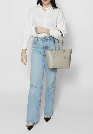 Vrouw met een wit overhemd, lichtblauwe spijkerbroek, zwarte puntige schoenen, met een beige boodschappentas over de schouder, staand tegen een grijze achtergrond.