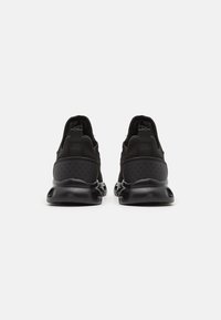 Zapatillas deportivas negras con parte superior texturizada, paneles perforados y suelas de goma con recortes. Diseñadas sin cordones, presentando un estilo minimalista.
