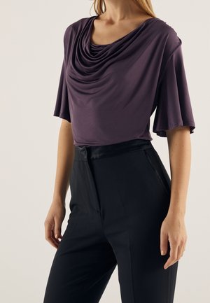 Femme portant un haut violet drapé à col bénitier avec des manches aux coudes et un pantalon noir taille haute sur un fond uni.