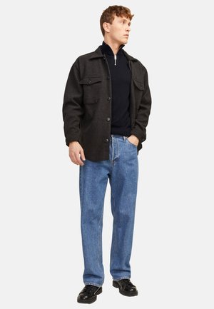 Junger Mann steht, trägt ein schwarzes, geknöpftes Overshirt, ein schwarzes Oberteil mit Reißverschluss, blaue Jeans und schwarze Schuhe, mit einer Hand in der Tasche.