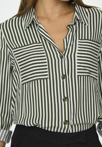 Chemise boutonnée à rayures vertes et blanches avec deux poches poitrine, col classique et manches retroussées. Le tissu semble léger et texturé.