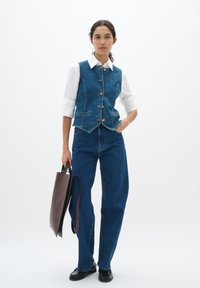 Donzen vest in donkerblauw met messing knopen en twee voorzakken, gecombineerd met hooggeknoopte blauwe jeans en een donkerbruine tote bag.