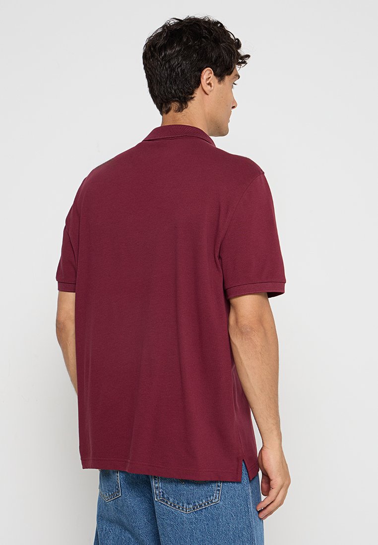 Polo rouge bourgogne à manches courtes, col plat, fentes latérales et tissu texturé. Le design est décontracté et légèrement surdimensionné.