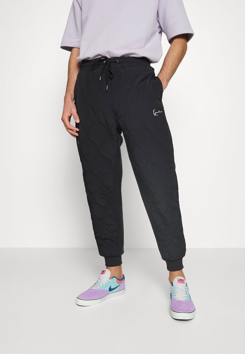 Homme portant un pantalon de jogging noir matelassé avec taille à cordon de serrage et des baskets multicolores, debout avec une main dans la poche.