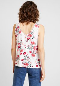 Donna con capelli ricci castani, che indossa un top senza maniche bianco con stampa floreale rossa e rosa e jeans blu, voltata di spalle rispetto alla telecamera.