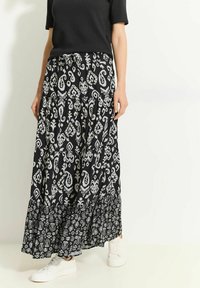 Jupe maxi noire à motifs avec dessins paisley blancs et formes géométriques, dotée d'un ourlet à volants et d'une taille élastique, portée avec des baskets blanches.